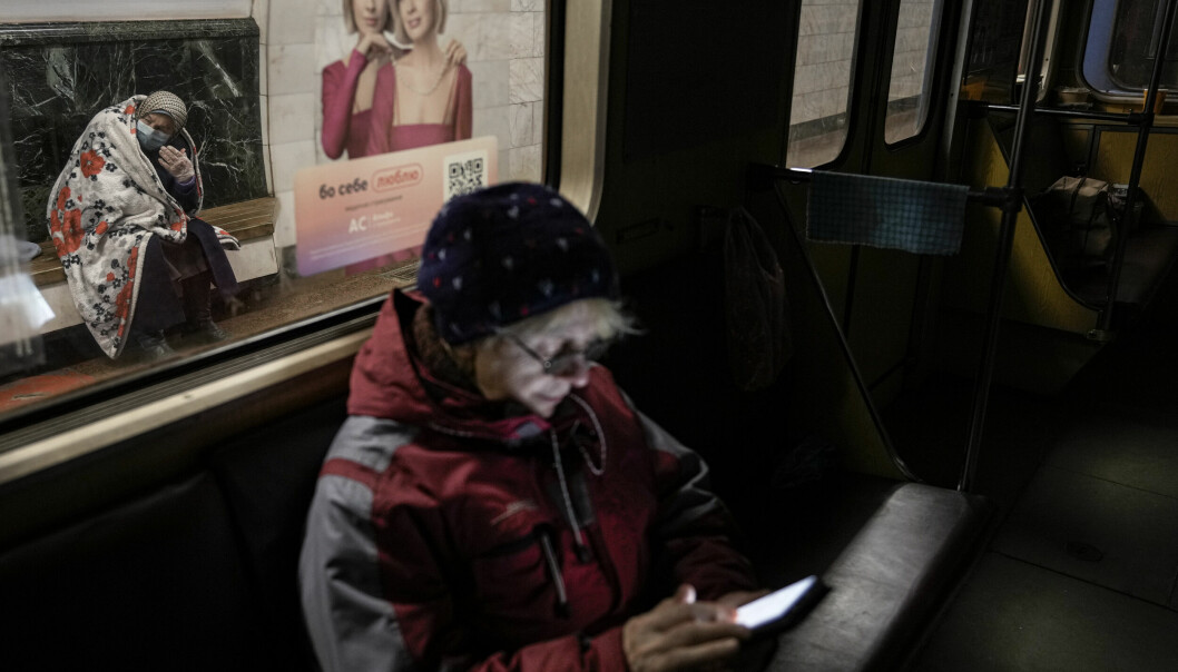 En äldre kvinna sitter med en filt i bakgrunden och i förgrunden en kvinna med en telefon. Tunnelbanestation i Kiev. TT / AP